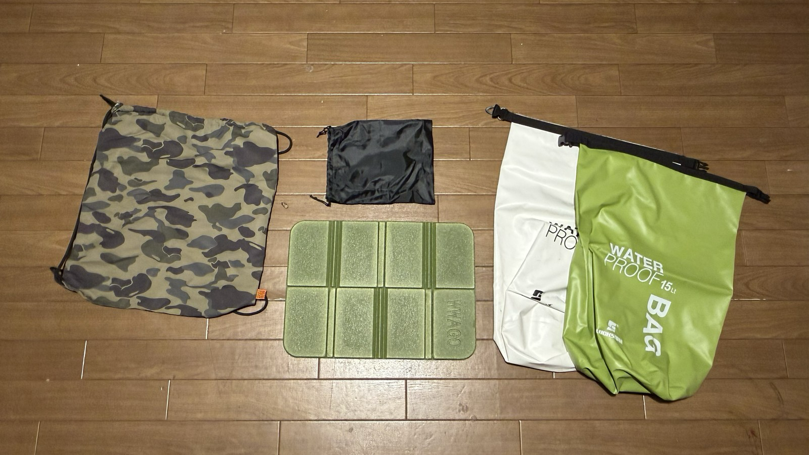 温泉に行く時に使う迷彩柄の巾着バッグ、サウナ用のお尻に敷くパッド(黒い収納袋と広げた状態)、白と緑の防水バッグを並べた写真